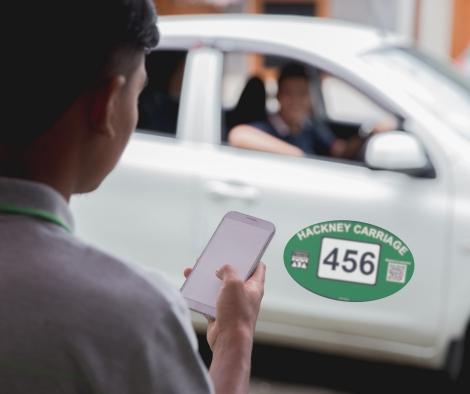 A hackney carriage vehicle with sign on door and someone with a mobile phone standing nearby