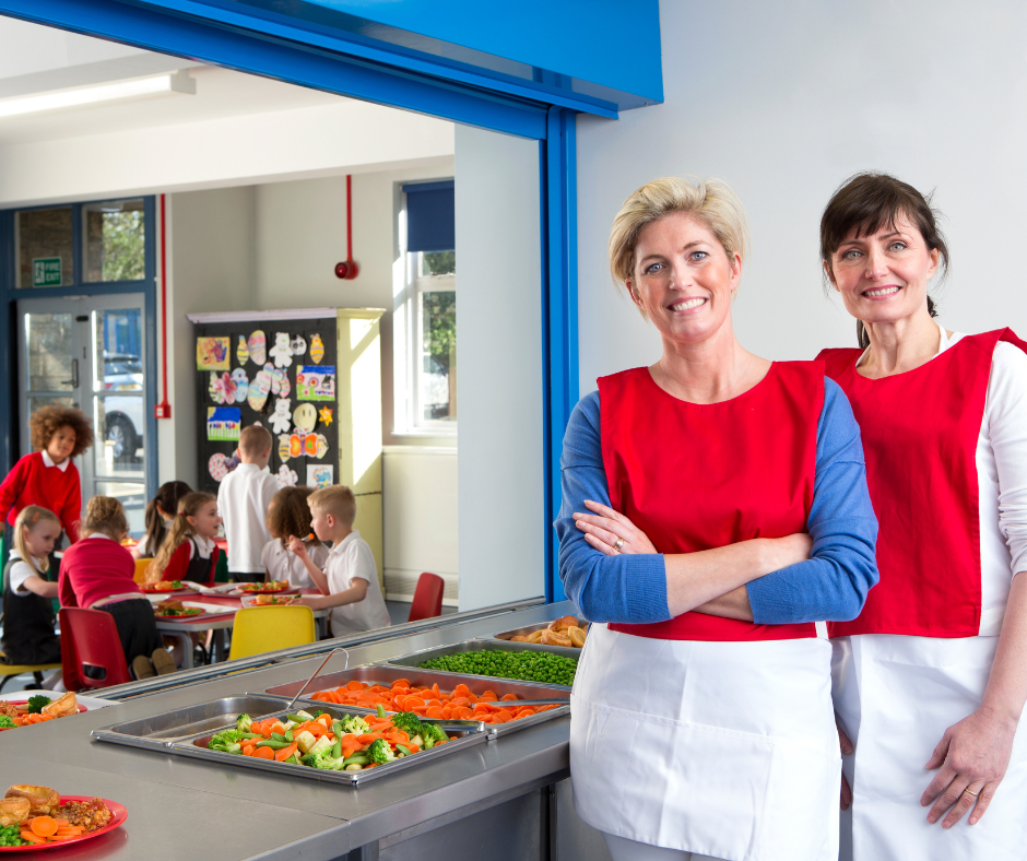 Two female school dinner staff with children eating in background.